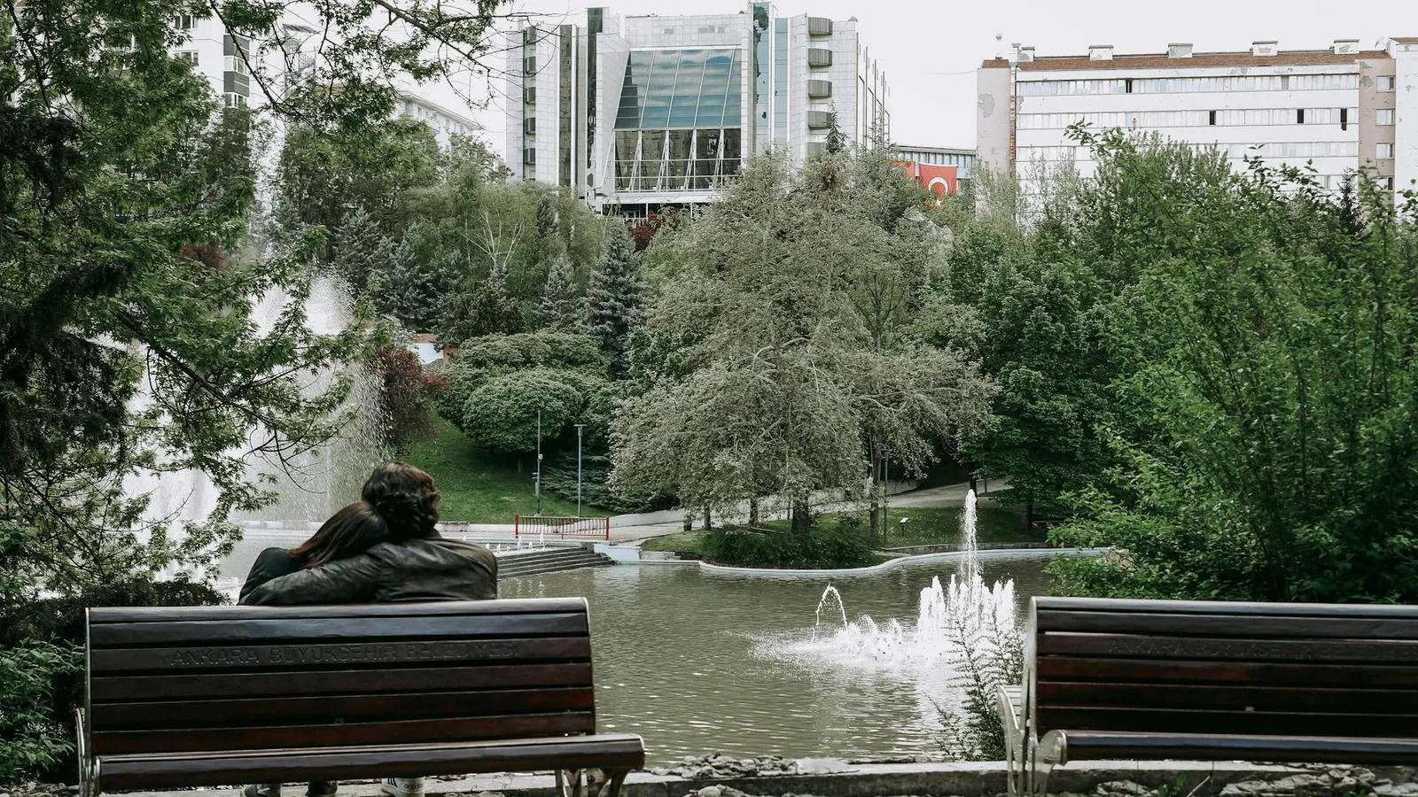 A couple relaxes on a bench overlooking a peaceful fountain and greenery in a city park setting.