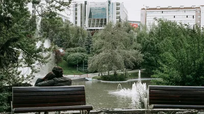 A couple relaxes on a bench overlooking a peaceful fountain and greenery in a city park setting.