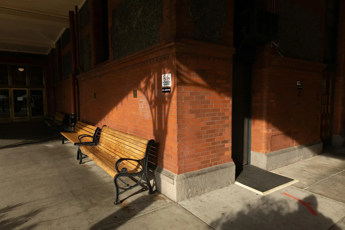 A street scene showing wooden benches against a brick facade under bright sunlight casting shadows.