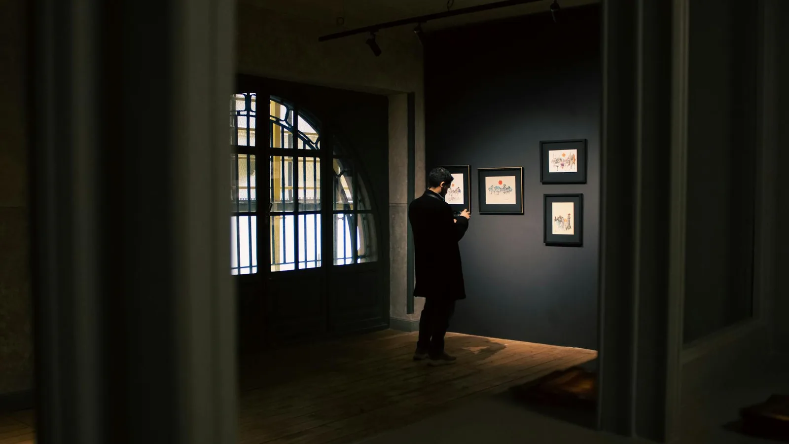 A man in a gallery analyzes framed art in dim lighting, creating a sophisticated atmosphere.