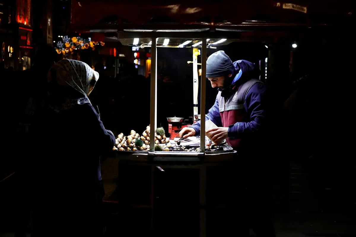 A street vendor selling food at night from a lit cart in an urban setting, interacting with a customer.