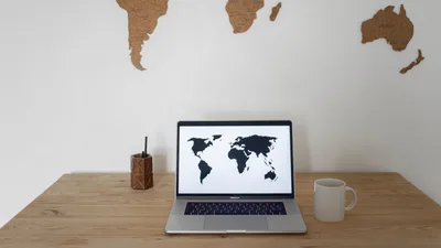 Black world map on laptop screen and ceramic cup with pen container placed on table against silhouettes of continents