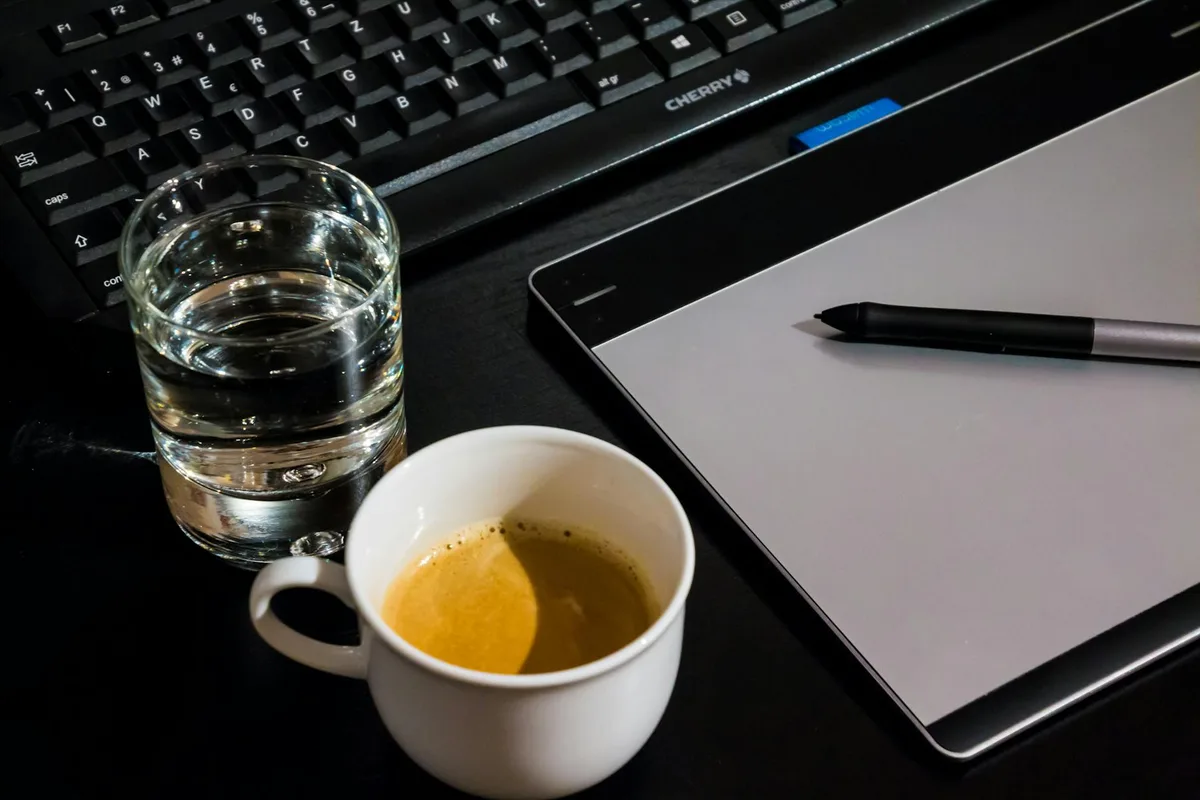 Office desk setup with a coffee cup, glass of water, tablet, pen, and keyboard.
