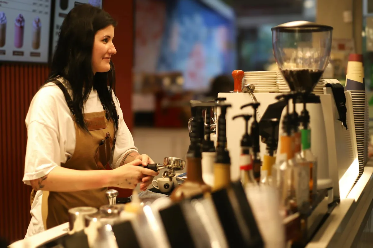 Barista in an apron skillfully making coffee in a bustling café environment with a variety of syrups and equipment.
